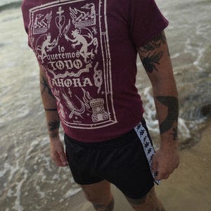 Puede incluir: Una camiseta color granate con un estampado gráfico blanco de un diseño que incluye un corazón, llamas y el texto "lo queremos todo ahora". La persona lleva pantalones cortos negros con una raya blanca a los lados.