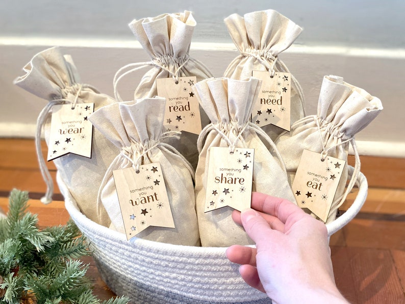 May include: A white wicker basket filled with small white fabric bags. Each bag has a wooden tag attached with a gold star design and text that reads "something you wear", "something you read", "something you need", "something you want", "something you share", and "something you eat".