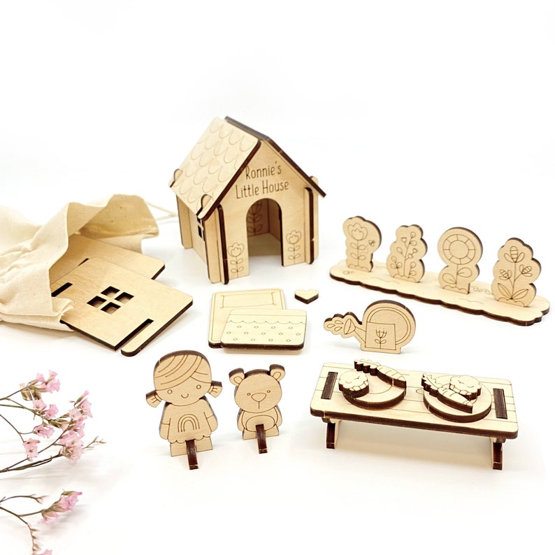 May include: A wooden playset for children, featuring a house, a bed, a table, a watering can, flowers, a girl doll, and a bear doll. The house has the text "Ronnie's Little House" on the front.