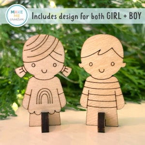 May include: Two wooden cutout figures of a girl and a boy, both with smiling faces. The girl has a rainbow on her shirt and the boy has a striped shirt. The figures are standing on small wooden bases. The text "Includes design for both GIRL + BOY" is visible above the figures.