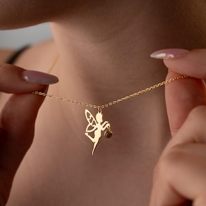 May include: A gold chain necklace with a delicate fairy pendant. The fairy is holding a small, round gemstone.
