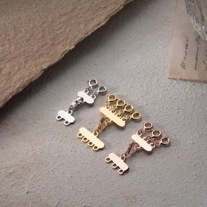 May include: Three silver, gold, and rose gold metal jewelry clasps with multiple loops. The clasps are designed to connect multiple strands of jewelry.
