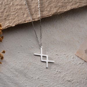 May include: A silver chain necklace with a geometric pendant in the shape of a cross. The pendant is made of a single piece of metal and has a simple, modern design.