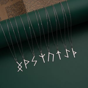 May include: Ten silver necklaces with different rune symbols hanging from them. The necklaces are arranged in a row on a green surface.