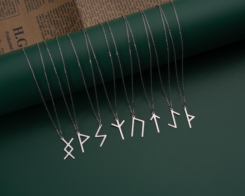 May include: Ten silver necklaces with different rune symbols hanging from them. The necklaces are arranged in a row on a green surface.