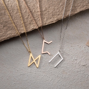 May include: Four necklaces with different geometric pendant designs. The pendants are made of gold, silver, and rose gold metal. The necklaces are hanging on a brown textured surface.