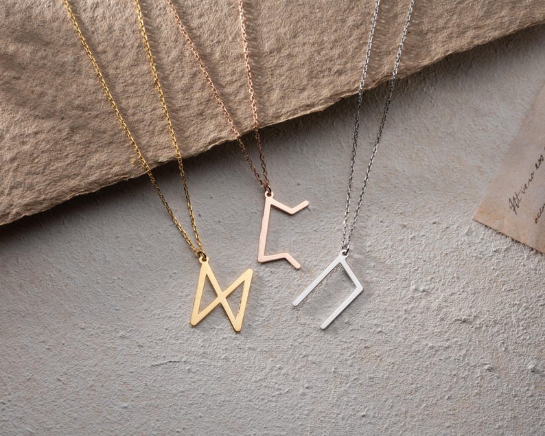 May include: Four necklaces with different geometric pendant designs. The pendants are made of gold, silver, and rose gold metal. The necklaces are hanging on a brown textured surface.