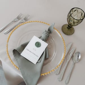 Può includere: Elegante mise en place con un piatto di vetro trasparente con bordo di perline dorate, un tovagliolo grigio piegato e un segnaposto. Le posate d'argento includono forchette, coltelli e un cucchiaio. Un bicchiere verde e una tovaglia grigio chiaro completano l'allestimento.