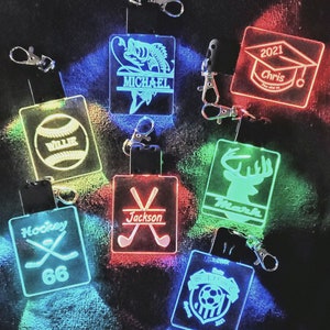 May include: Seven different personalized LED keychains with various sports and hobbies themes. The keychains are illuminated in different colors, including red, green, blue, and yellow. The keychains feature names like "Michael", "Chris", "Mark", "Jackson", "Willie", and "66".