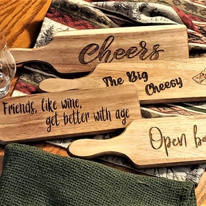 May include: Four wooden serving boards with engraved sayings. The sayings are "Cheers", "The Big Cheesy", "Friends, like wine, get better with age", and "Open bar".