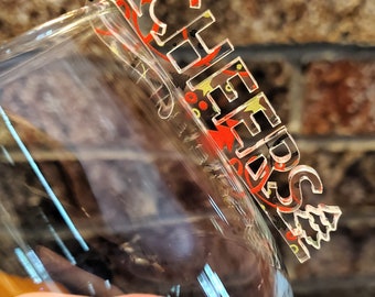May include: A clear glass wine glass with a red, yellow, and black acrylic "Cheers!" charm attached to the rim.