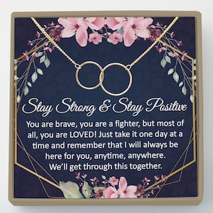 May include: A gold-colored necklace with two interlocking circles, presented on a navy blue background with floral accents. The text "Stay Strong & Stay Positive" is displayed above a supportive message.