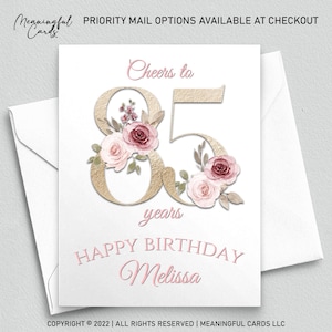 May include: A white birthday card with a gold textured "85" and floral accents in pink and burgundy. The card reads "Cheers to 85 years" and "Happy Birthday Melissa" in pink script. Includes a white envelope.