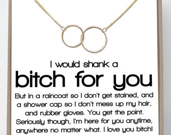 Puede incluir: Un collar dorado con dos círculos entrelazados, presentado en una tarjeta blanca con un mensaje humorístico. El texto dice "I would shank a bitch for you" con texto adicional debajo. La tarjeta está enmarcada.
