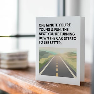 May include: A white card with black text that reads "One minute you're young & fun. The next you're turning down the car stereo to see better." The card has a picture of a road with a sunset in the background.