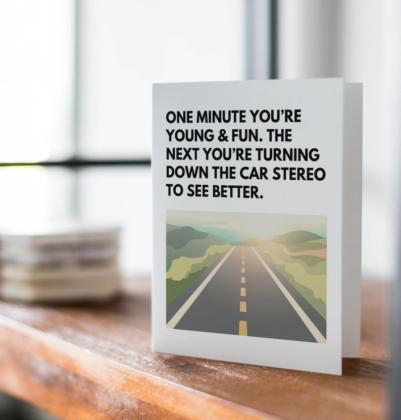 May include: A white card with black text that reads "One minute you're young & fun. The next you're turning down the car stereo to see better." The card has a picture of a road with a sunset in the background.