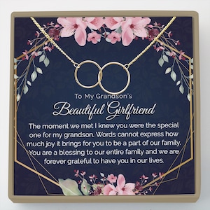 May include: A gold-colored necklace with two interlocking circles, presented on a navy blue card with floral accents. The card reads "To My Grandson's Beautiful Girlfriend" with a heartfelt message.