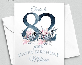 May include: A white birthday greeting card featuring the large dark blue watercolor number 82 decorated with pink flowers and dark blue leaves. The text reads Cheers to 82 years HAPPY BIRTHDAY Melissa in blue script and block lettering.