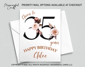 May include: A white card with a black number 35 decorated with pink and brown floral illustrations. The text "Cheers to" is above the number and "years" is below the number. The text "HAPPY BIRTHDAY Chloe" is below the number.