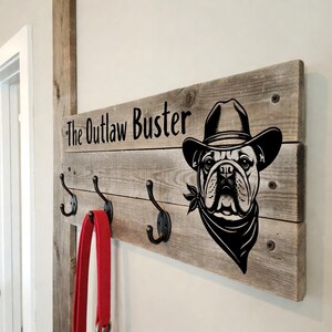 Puede incluir: Un perchero de pared rústico de madera con cuatro ganchos negros. Presenta un gráfico negro de un bulldog con sombrero de vaquero y pañuelo, con el texto "The Outlaw Buster". Una correa roja cuelga de un gancho.