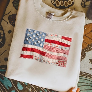 Peut inclure: Sweat-shirt crème or beige or crème clair or beige clair or blanc cassé or blanc crème avec un drapeau américain en patchwork. Le drapeau comprend des étoiles bleues, des rayures rouges et blanches et des motifs floraux et à carreaux. Col rond.