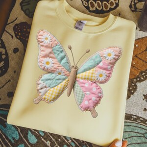 Peut inclure: Un sweat-shirt jaune pâle orné d'un grand papillon en patchwork. Le papillon a des ailes aux motifs roses pastel, bleus et jaunes à carreaux et à pois, avec des accents de marguerites blanches. Le corps du papillon est de couleur beige.