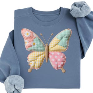 Peut inclure: Un sweat-shirt bleu avec un motif de papillon. Les ailes du papillon sont en patchwork avec des motifs floraux, à carreaux et à pois en rose, jaune et vert. Le sweat-shirt a un col rond et des manches longues.