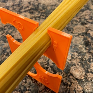 May include: A bright orange plastic spaghetti measuring tool with a cross-shaped design holding a bundle of dry yellow spaghetti noodles. The tool features embossed icons including two interlocking circles and a small ice cream cone shape on its surface.