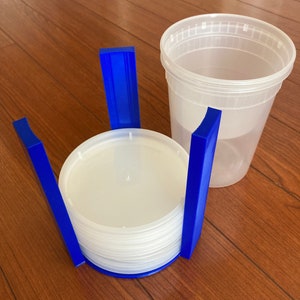 May include: A blue plastic storage container organizer with a stack of clear plastic lids inside. The organizer has four vertical supports and is designed to hold lids upright.