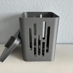 May include: A dark gray, square-shaped container with a textured finish and a matching lid placed to the side. The body of the container features vertical, elongated cutouts on its sides and a star-shaped opening at the base for ventilation.