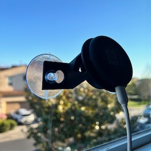 May include: A black car phone mount with a suction cup and a clear plastic disc. The mount is attached to a window and a white cable is connected to the phone mount.