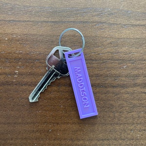 May include: A purple keychain with the name "MADDISON" engraved on it. A silver key is attached to the keychain.