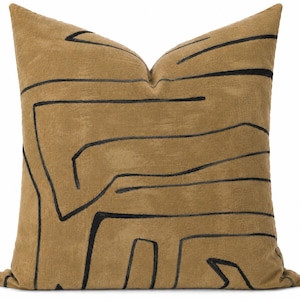 May include: A square throw pillow in a muted tan color featuring a bold, black embroidered line pattern that creates an abstract, interconnected design across the front surface. The pillow has a soft, textured appearance with a visible seam.