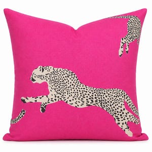 May include: A square throw pillow in a vibrant hot pink color. The pillow features a repeating pattern of two cheetahs in a leaping motion, rendered in a cream color with black spots. The fabric has a visible woven texture throughout the surface.