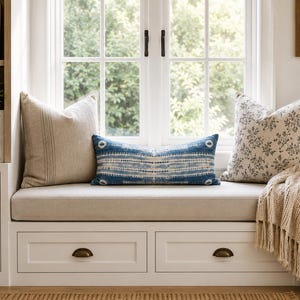 Puede incluir: Un banco de ventana blanco con dos cajones de almacenamiento con tiradores de lat&oacute;n, cubierto con un coj&iacute;n beige. Tres cojines est&aacute;n dispuestos en el asiento: uno cuadrado beige liso, uno lumbar con patr&oacute;n azul y blanco y uno floral.