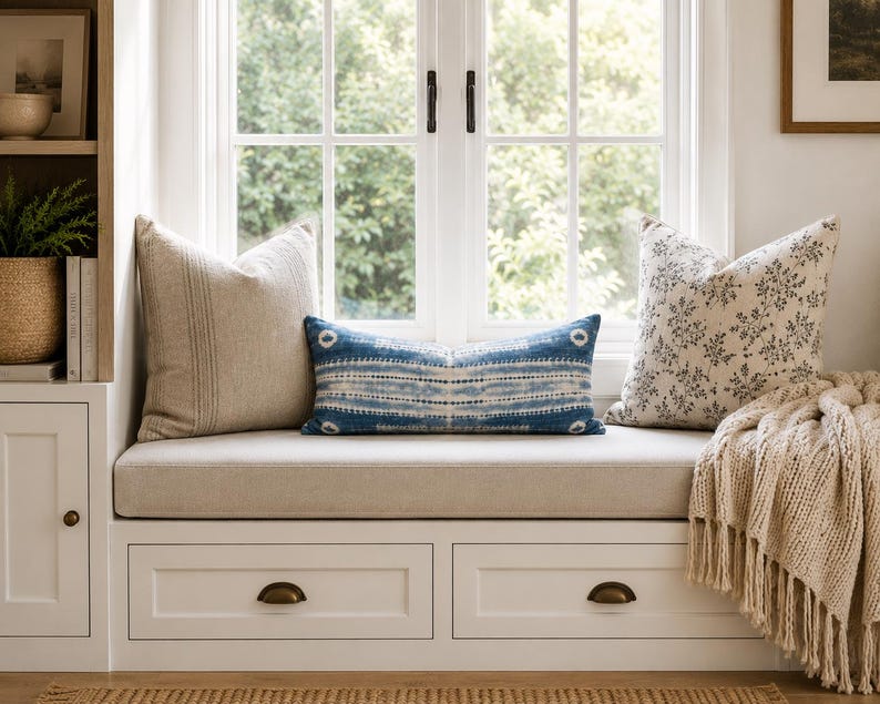 Puede incluir: Un banco de ventana blanco con dos cajones de almacenamiento con tiradores de lat&oacute;n, cubierto con un coj&iacute;n beige. Tres cojines est&aacute;n dispuestos en el asiento: uno cuadrado beige liso, uno lumbar con patr&oacute;n azul y blanco y uno floral.