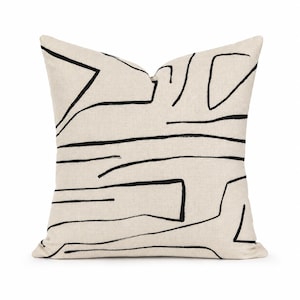 May include: A square throw pillow featuring a light beige, textured fabric base. The surface is decorated with a series of bold, black hand-drawn lines that form abstract, geometric shapes and irregular patterns across the entire front face.