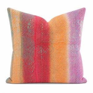 May include: A square throw pillow featuring a textured, nubby surface with a vertical gradient pattern. The color palette transitions through shades of orange, vibrant red, soft yellow, and purple, with subtle hints of teal woven throughout the fabric.