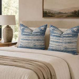 Puede incluir: Dos cojines cuadrados decorativos con un patr&oacute;n azul y blanco estilo te&ntilde;ido anudado, con l&iacute;neas horizontales discontinuas y peque&ntilde;os motivos circulares. Los cojines est&aacute;n colocados sobre una cama con ropa de cama y cabecero beige.