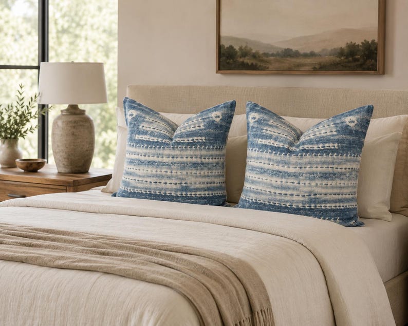 Puede incluir: Dos cojines cuadrados decorativos con un patr&oacute;n azul y blanco estilo te&ntilde;ido anudado, con l&iacute;neas horizontales discontinuas y peque&ntilde;os motivos circulares. Los cojines est&aacute;n colocados sobre una cama con ropa de cama y cabecero beige.