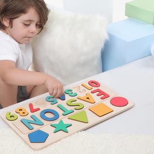 May include: A wooden puzzle with the name "EASTON" in colorful letters and numbers, along with various shapes. The puzzle is on a white surface, with a child interacting with it. Additional loose numbers are nearby.