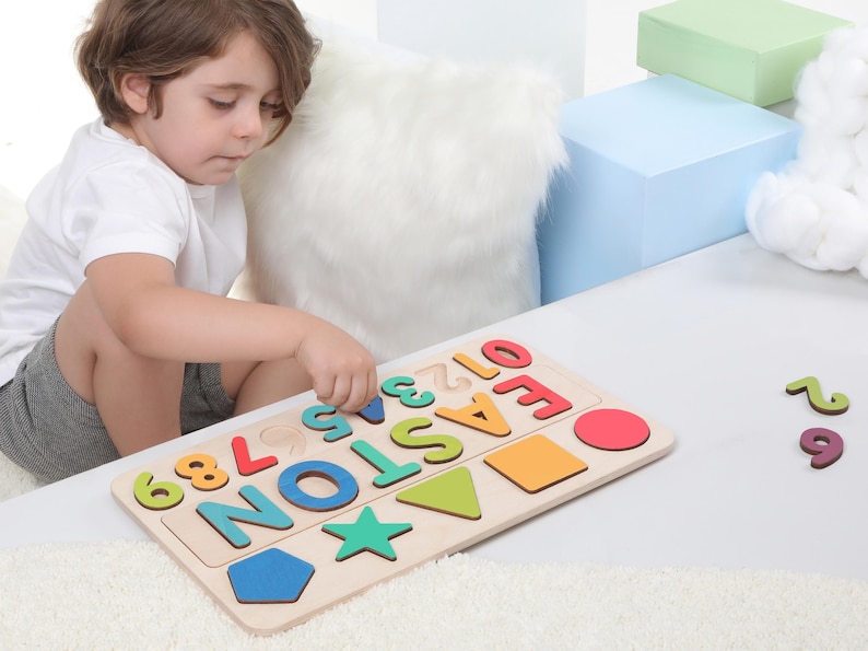 May include: A wooden puzzle with the name "EASTON" in colorful letters and numbers, along with various shapes. The puzzle is on a white surface, with a child interacting with it. Additional loose numbers are nearby.