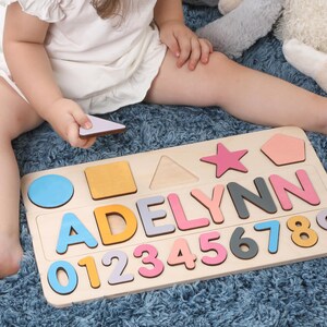 May include: Wooden puzzle board with the name "ADELYNN" spelled out in colorful letters. Includes number and shape cutouts. The puzzle is on a light wood base, with a child holding a puzzle piece.