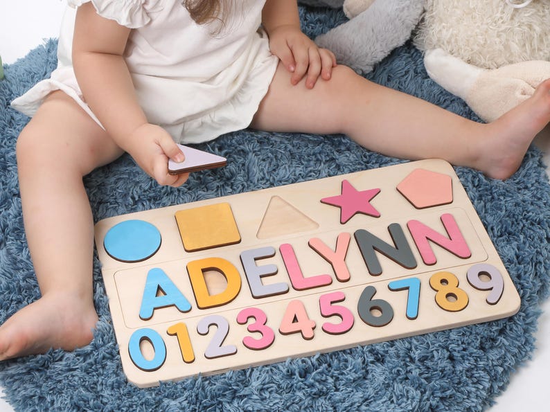May include: Wooden puzzle board with the name "ADELYNN" spelled out in colorful letters. Includes number and shape cutouts. The puzzle is on a light wood base, with a child holding a puzzle piece.