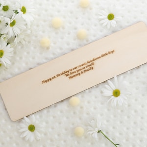 May include: A rectangular wooden plaque with engraved text, surrounded by white daisies and small yellow pom-poms. The text reads "Happy 1st Birthday to our sweet, handsome little boy! We love you so much! Mommy & Daddy."