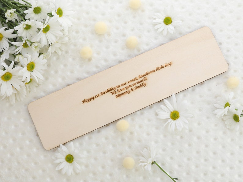 May include: A rectangular wooden plaque with engraved text, surrounded by white daisies and small yellow pom-poms. The text reads "Happy 1st Birthday to our sweet, handsome little boy! We love you so much! Mommy & Daddy."
