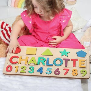 May include: Wooden name puzzle with the name "CHARLOTTE" in colorful letters and numbers 0-9. The puzzle also includes various shapes. The child is wearing a pink dress and is interacting with the puzzle.