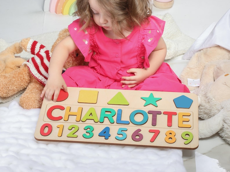 May include: Wooden name puzzle with the name "CHARLOTTE" in colorful letters and numbers 0-9. The puzzle also includes various shapes. The child is wearing a pink dress and is interacting with the puzzle.