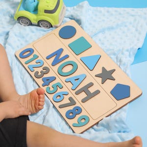 May include: Wooden puzzle board with the name "NOAH" in blue and gray letters, along with numbers and geometric shapes. A green and blue toy car and a blue toy car are also in the image. The puzzle is on a light blue blanket.
