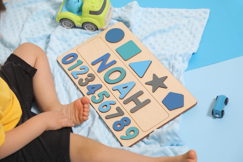 May include: Wooden puzzle board with the name "NOAH" in blue and gray letters, along with numbers and geometric shapes. A green and blue toy car and a blue toy car are also in the image. The puzzle is on a light blue blanket.